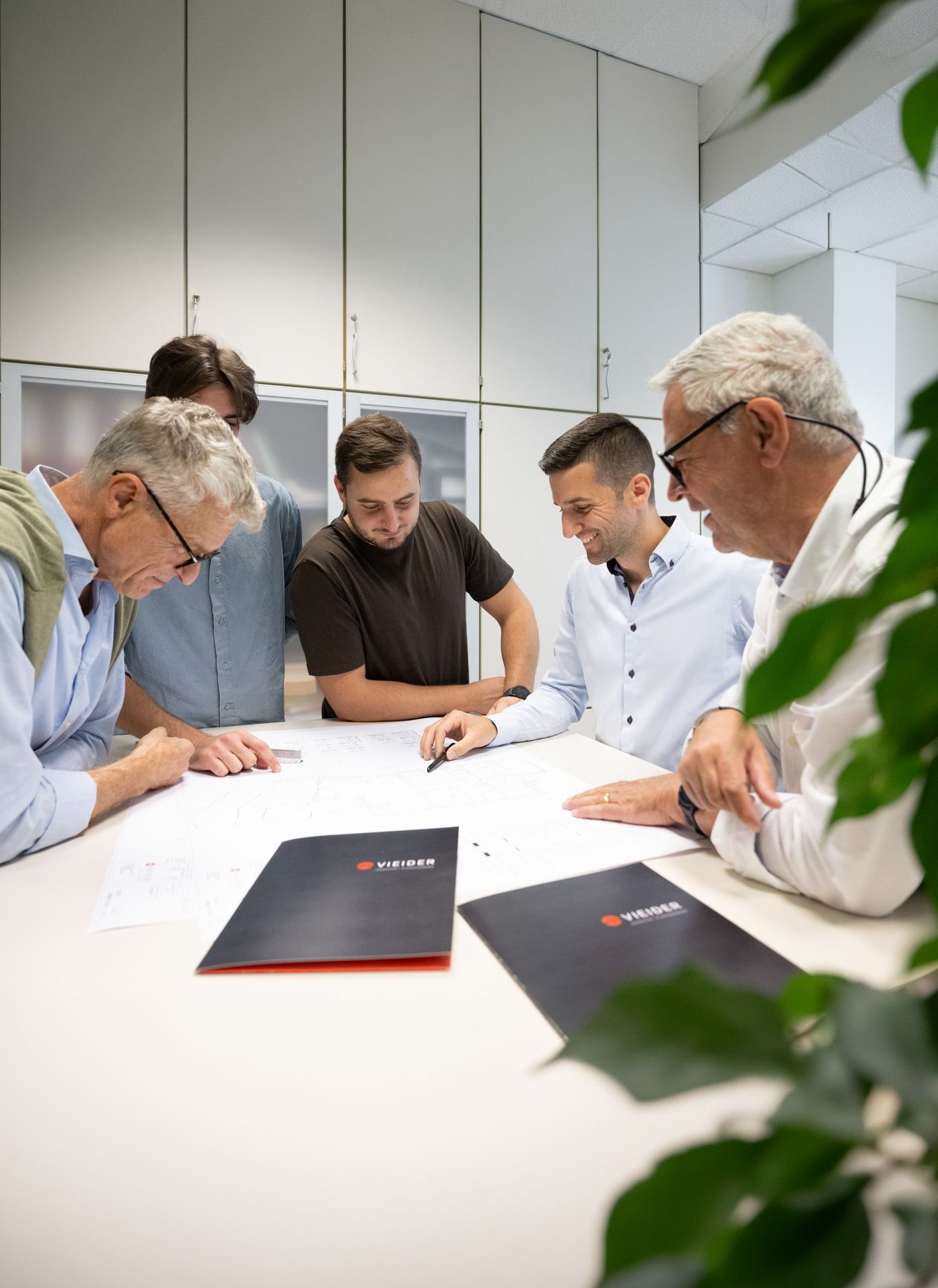 Fünf Männer besprechen an einem Tisch technische Zeichnungen und Unterlagen in einem hellen Büro.