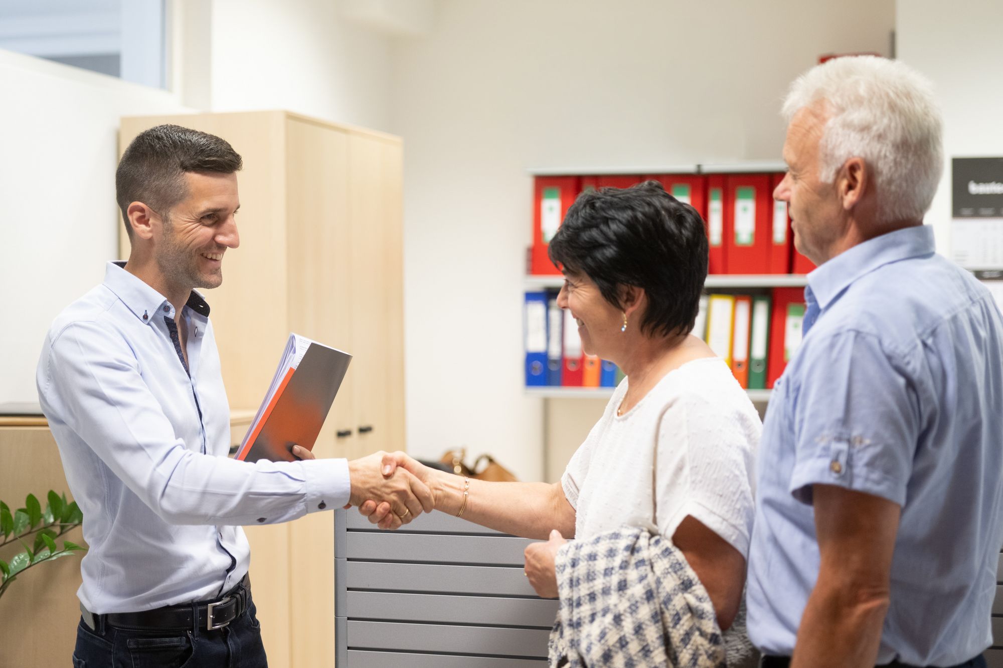 Ein lächelnder Mann mit Unterlagen schüttelt einer Frau in einem Büro die Hand, während ein älterer Mann danebensteht.