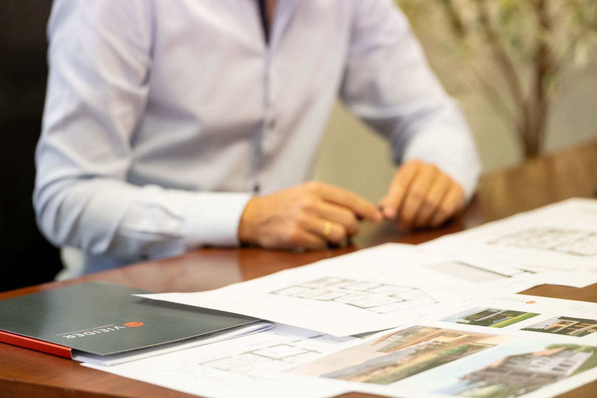 Person im weißen Hemd arbeitet an einem Tisch mit Bauplänen, Architekturunterlagen und einem Katalog.
