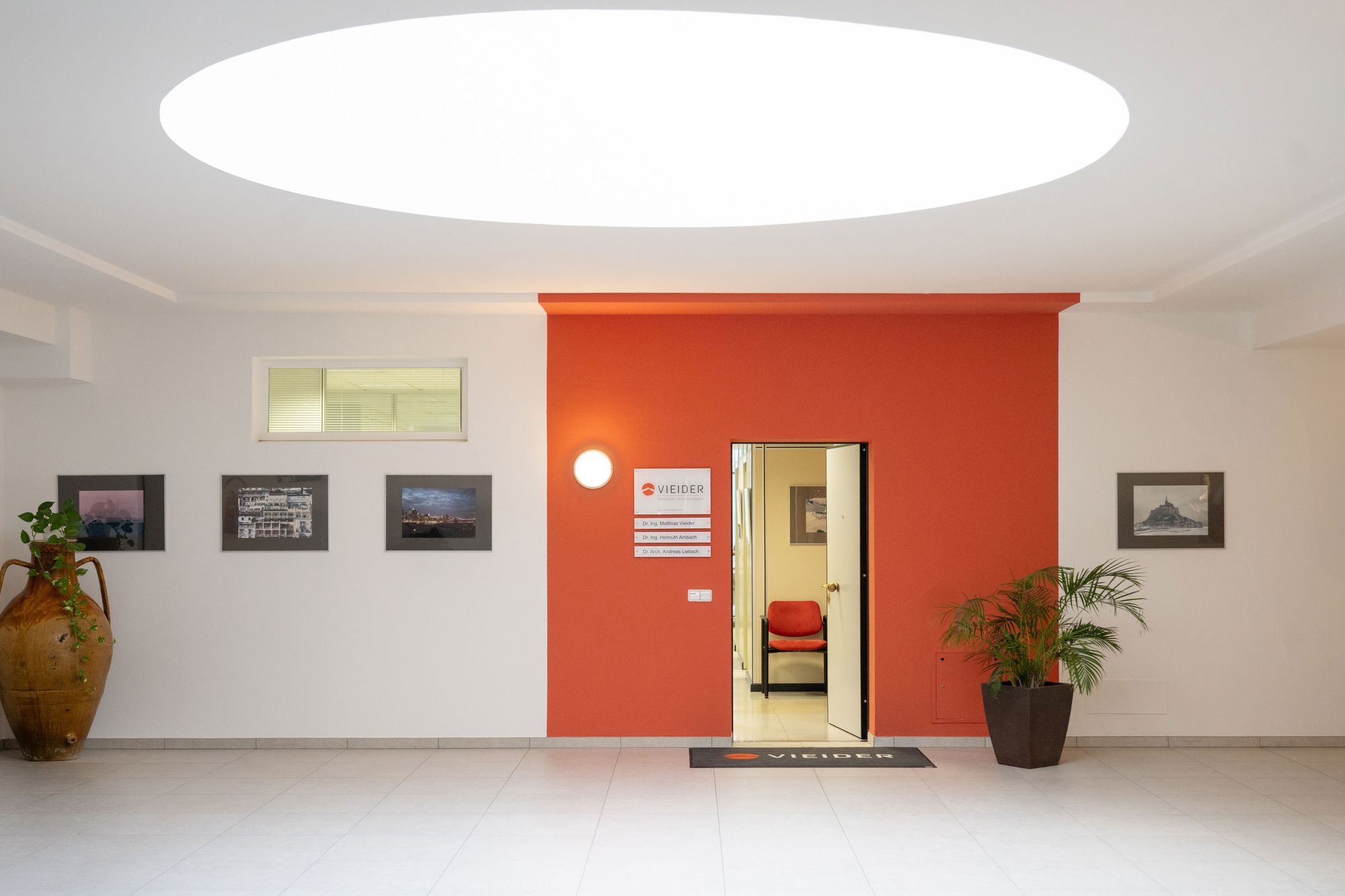 Heller Empfangsbereich mit roter Akzentwand, geöffneter Tür, gerahmten Bildern, Pflanzen und großem ovalem Deckenlicht.