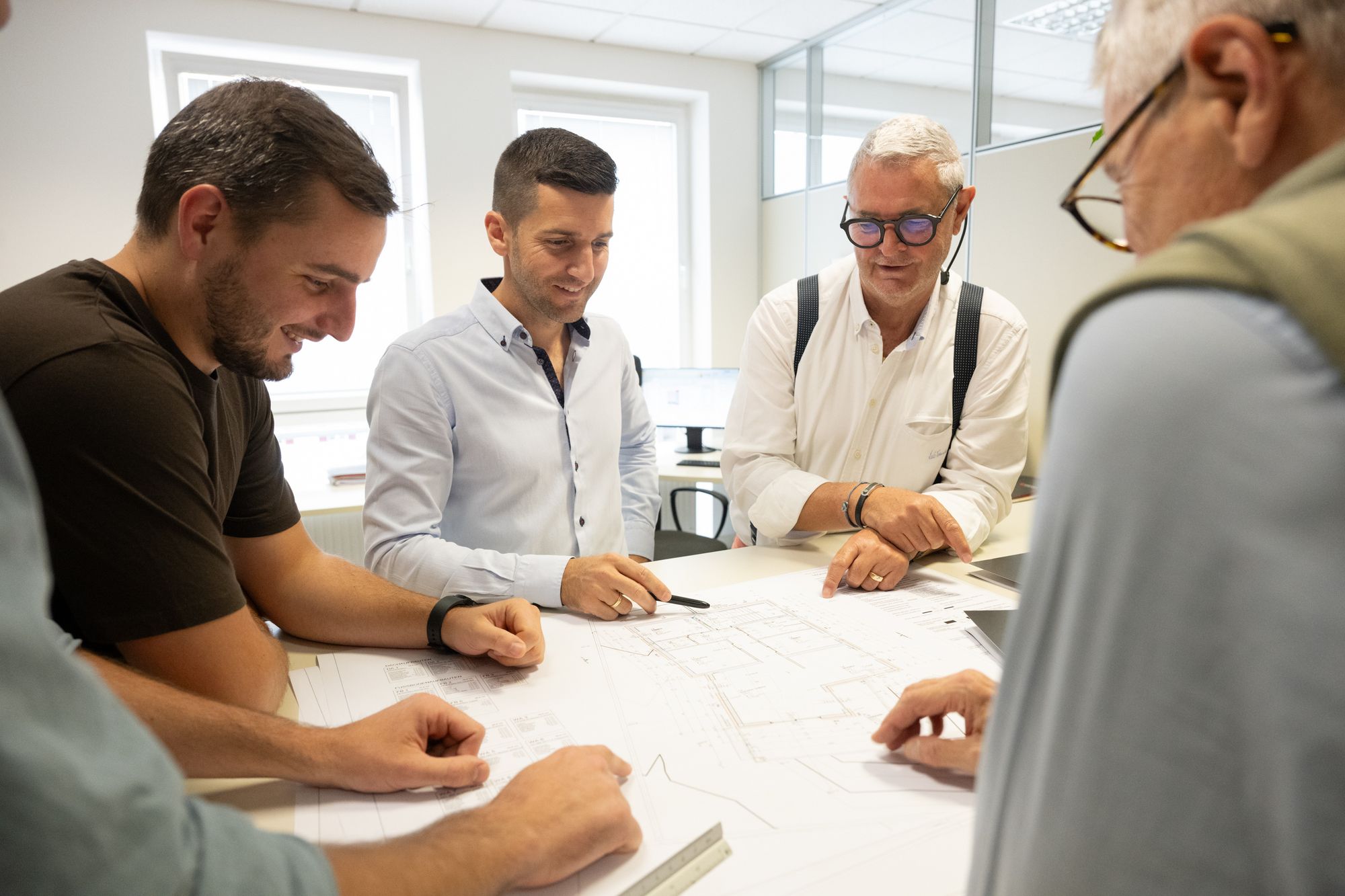 Mehrere Männer besprechen gemeinsam technische Zeichnungen an einem Tisch in einem hellen Büro.