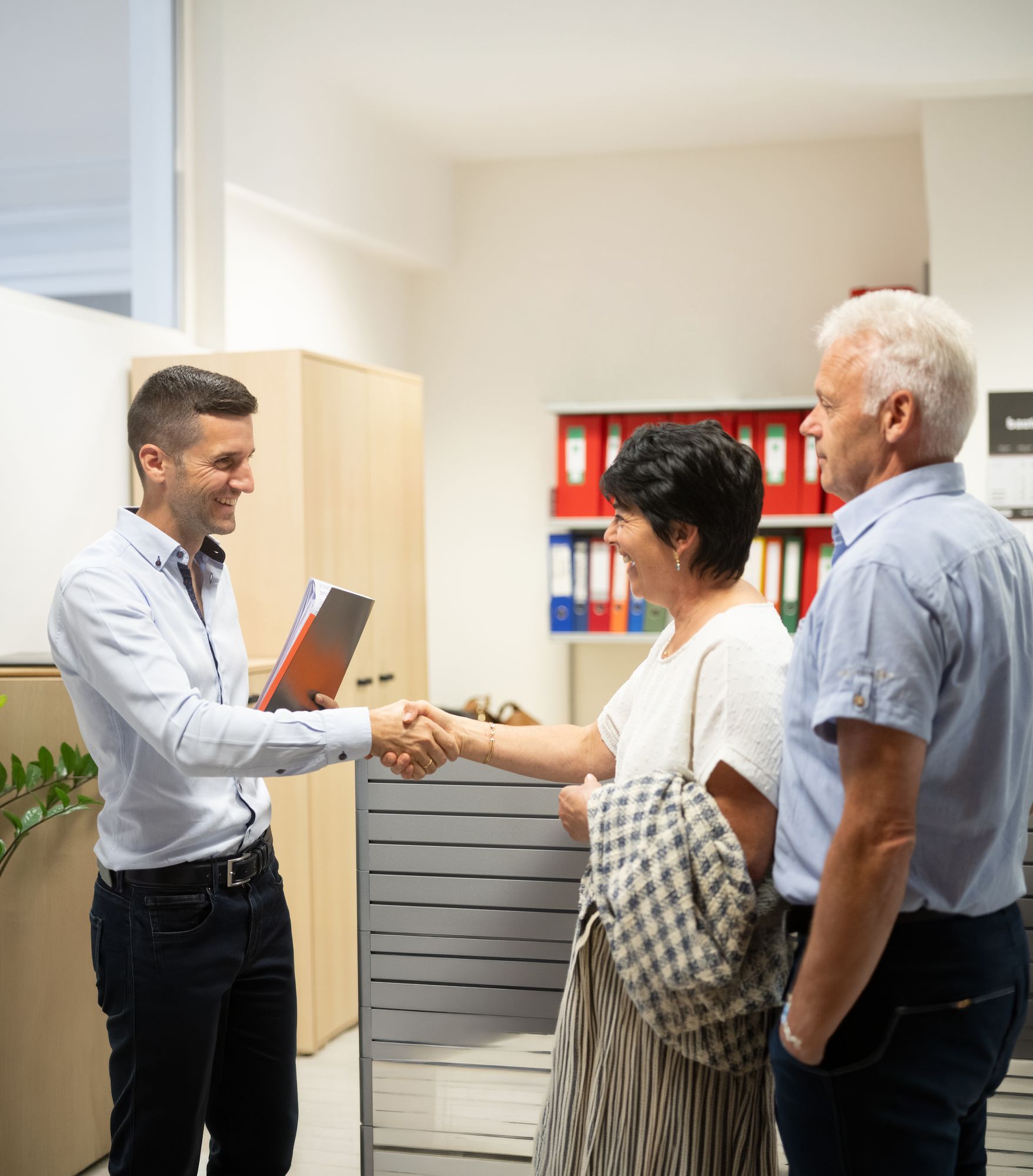 Ein Mann begrüßt ein älteres Ehepaar per Handschlag in einem Büro mit bunten Aktenordnern.